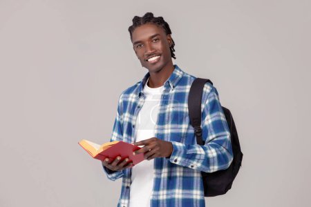 Foto de Concepto educativo. Guapo chico afroamericano con mochila y libro posando aislado sobre fondo gris estudio, feliz estudiante negro macho sonriendo y mirando a la cámara, espacio de copia - Imagen libre de derechos