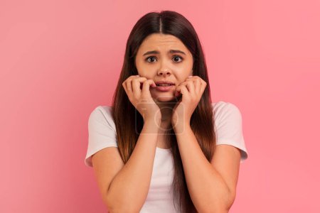 Stressed teen girl touching face with hands and looking at camera, scared emotional female teenager standing against pink studio background, suffering moment of panic or concern, copy space