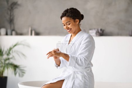 Téléchargez les photos : Souriant noir femelle appliquant crème pour les mains tout en étant assis sur la baignoire dans la salle de bain moderne à la maison, Happy African American Woman portant peignoir blanc faisant des traitements de beauté, Copier l'espace - en image libre de droit