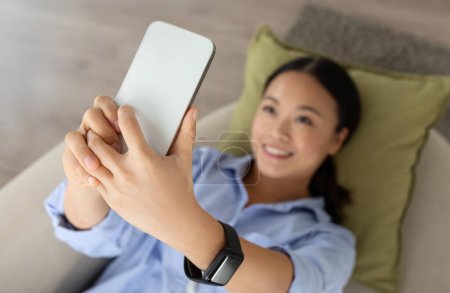 Photo for Selective focus on modern smartphone in asian woman hands, home interior. Happy korean lady resting on sofa, taking selfie on cell phone, have video call with lover, smiling at phone camera - Royalty Free Image