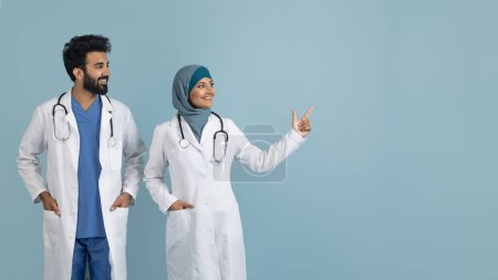Téléchargez les photos : Souriant arabe musulman professionnels de la santé en manteaux blancs pointant vers l'espace de copie à droite, heureux jeune couple de médecins du Moyen-Orient debout sur fond bleu studio, panorama - en image libre de droit