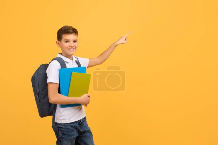 Téléchargez les photos : Offre éducative. Souriant adolescent garçon tenant des livres et pointant du doigt à part à l'espace de copie, heureux écolier avec sac à dos et cahiers indiquant endroit gratuit pour la publicité sur fond jaune - en image libre de droit