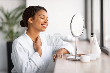 Foto de Hermosa mujer negra mirando en el espejo y tocando la piel del cuello, sonriente mujer afroamericana en albornoz blanco comprobando arrugas en la zona de decollete, disfrutando de tratamientos de belleza domésticos, primer plano - Imagen libre de derechos