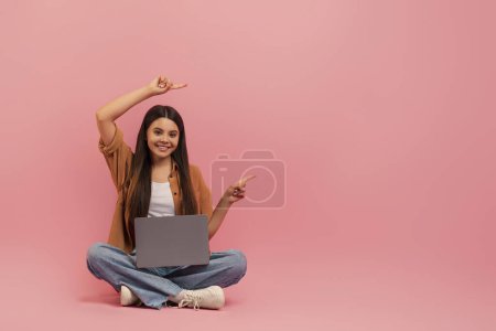 Téléchargez les photos : Adolescente joyeuse assise sur le sol avec ordinateur portable et pointant du doigt avec deux mains, heureuse adolescente démontrant l'espace de copie pour la publicité ou l'offre, posant sur fond de studio rose - en image libre de droit