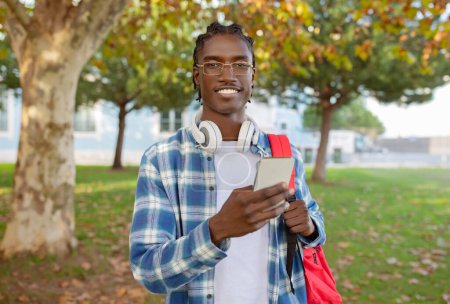 Photo for African American student guy casually scrolling through social media on his phone, texting and communicating holding smartphone, standing with headphones and backpack outdoors - Royalty Free Image
