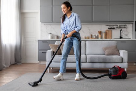 Photo for Focused and diligent, young woman vacuums the living room carpet, wielding modern vacuum cleaner with ease as she maintains the cleanliness of her home - Royalty Free Image