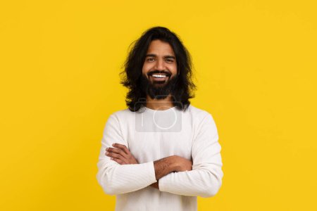 Téléchargez les photos : Élégant positif beau jeune Indien barbu aux cheveux longs portant une tenue blanche posant isolé sur fond de studio jaune, tenant les bras croisés sur la poitrine, souriant à la caméra, espace de copie - en image libre de droit