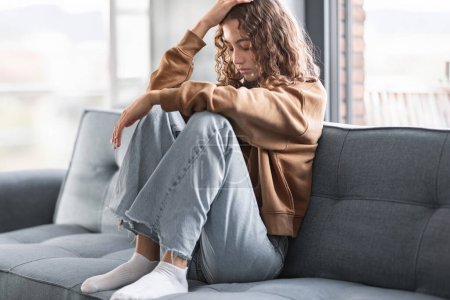 Photo for Teenage depression. Frustrated European adolescent girl touches head in moment of loneliness and sadness, reflecting concept of youth mental health challenges, while seated on sofa indoor - Royalty Free Image