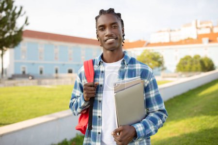 Téléchargez les photos : Un étudiant afro-américain avec un sac à dos contenant des cahiers d'exercices et un gadget pour ordinateur portable, se tient dans le parc du campus à l'extérieur, sourit à la caméra. Joyeux incarnation de la vie académique moderne - en image libre de droit