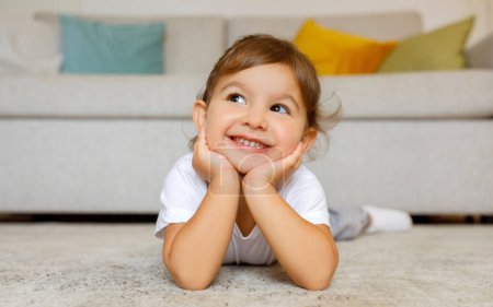Photo for Cheerful cute little girl daydreaming on the floor at home, adorable toddler female child lying on carpet in living room, resting head on hands, smiling and looking away, copy space - Royalty Free Image