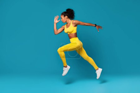 Photo for Athletic young black woman in yellow activewear leaping during dynamic workout on blue studio backdrop, epitome of strength and beauty of fitness. Full length, side view - Royalty Free Image