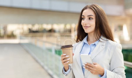 Photo for Multitasking happy caucasian businesswoman walking with confidence, holding a coffee cup and smartphone, wearing a formal suit, embodying urban professional lifestyle, panorama - Royalty Free Image