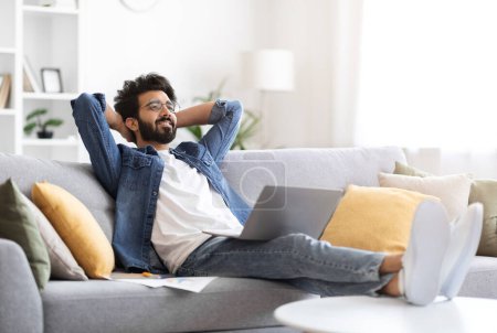 Téléchargez les photos : Heureux homme indien en tenue décontractée allongé sur le canapé avec son ordinateur portable, gai gars de l'Est reposant sur le canapé à la maison avec les mains derrière la tête, l'air satisfait, prendre une pause de travail à distance - en image libre de droit