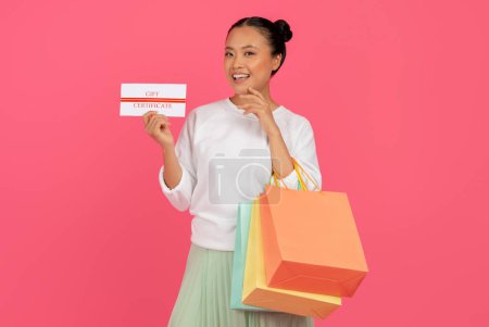 Téléchargez les photos : Femme asiatique excitée tenant des sacs à provisions et montrant un certificat-cadeau, Joyeux coréen femme a reçu bon de réduction après avoir fait des achats, Debout isolé sur fond de studio rose, Espace de copie - en image libre de droit