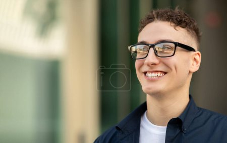 A smiling caucasian young man wearing glasses, a navy blue shirt, and a white t-shirt, with a blurred urban background look at free space to ad and offer, close up. Work, study