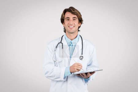 Smiling young male doctor in white lab coat holding digital tablet, ready to take notes or consult patients, epitomizing modern medical practice