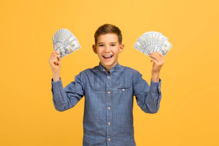 Téléchargez les photos : Jeune garçon excité tenant de l'argent comptant dans les deux mains, adolescent heureux garçon vêtu de chemise en denim debout sur fond jaune studio, illustrant la richesse, l'épargne ou le succès financier - en image libre de droit