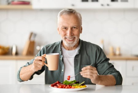 Hombre mayor sonriente almorzando y disfrutando de una taza de café, caballero mayor feliz desayunando, sentado en la mesa en la cocina y mirando a la cámara, mostrando una dieta equilibrada, espacio para copiar