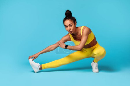 Photo for Athletic young black woman exercising during flexibility workout, stretching legs muscles, posing in extended leg squat position over blue studio backdrop, looking at camera. Sport, fitness - Royalty Free Image