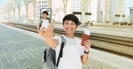 Photo for Happy young chinese guy travel blogger taking selfie on smartphone, showing passport and tickets, waiting for train. Cheerful asian millennial man tourist sharing photos on social media, banner - Royalty Free Image