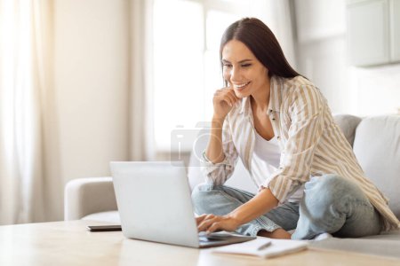 Photo for Smiling young woman using laptop for remote work at home, beautiful millennial lady sitting at coffee table in bright living room, exemplifying comfortable work-from-home setting, copy space - Royalty Free Image