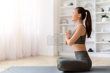 Foto de Tranquil joven mujer asiática meditando en casa, practicando yoga con los brazos cerrados, hermosa mujer coreana sentada en posición de oración en la estera en la sala de estar, irradiando paz interior, espacio para copiar - Imagen libre de derechos