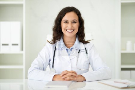 Photo for Friendly millennial caucasian woman doctor sitting at desk at white office interior at modern clinic, smiling at camera. Lady general practitioner have appointment with patient at hospital - Royalty Free Image