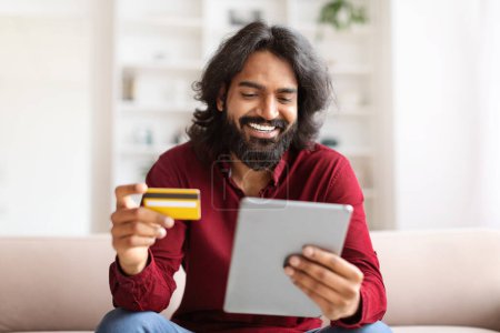 Photo for Happy indian guy enjoying newest shopping application, sitting on couch at home, using digital tablet and holding credit card, banking online, paying on Internet, e-commerce - Royalty Free Image
