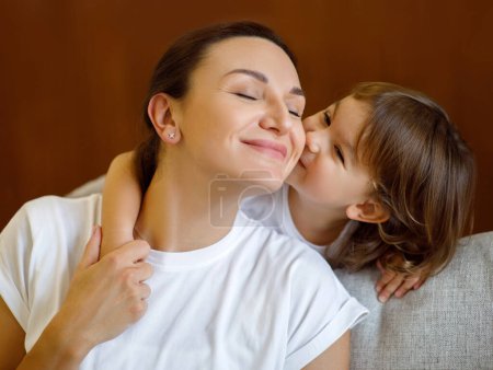 Photo for Mothers Day Concept. Cute Little Girl Kissing Her Mom In Cheek, Happy Young Mommy And Adorable Toddler Daughter Bonding Together At Home, Enjoying Tender Moments, Relaxing On Couch In Living Room - Royalty Free Image