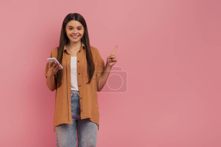 Photo for Happy teenage girl holding smartphone and pointing upwards with one hand, smiling teen female signifying good idea or promotion, standing against pink studio background, copy space - Royalty Free Image
