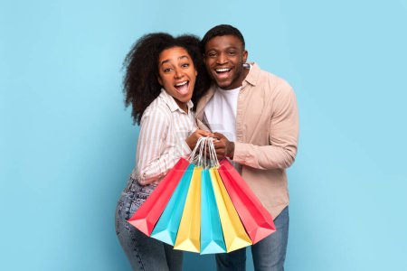 Photo for Radiant young african american couple sharing laugh, holding bunch of vibrant shopping bags, against refreshing blue backdrop - Royalty Free Image