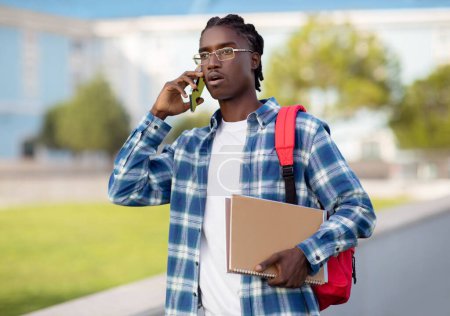 Photo for Shocked unhappy african american student guy talking on phone, solving educational college problem through mobile chat, walking with backpack and workbooks outdoors university campus - Royalty Free Image