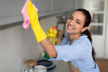 Foto de Mujer radiante caucásica en camisa azul y guantes amarillos, sosteniendo botella de spray, limpiando alegremente la superficie con tela rosa en una cocina bien iluminada - Imagen libre de derechos
