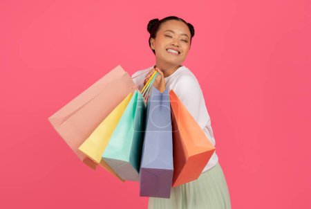 Photo for Cheerful Young Asian Shopaholic Woman Holding Bright Paper Shopping Bags, Happy Korean Female Carrying Purchases And Closing Eyes With Delight, Standing Isolated Over Pink Background, Copy Space - Royalty Free Image