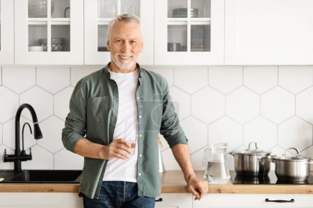 Téléchargez les photos : Homme mûr joyeux tenant un verre d'eau tout en posant dans la cuisine moderne, homme âgé heureux suggérant un mode de vie sain et l'hydratation, incarnant le bien-être et le contentement, l'espace de copie - en image libre de droit