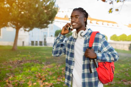 Téléchargez les photos : Un étudiant noir avec un sac à dos parlant sur un téléphone portable, bavardant en sortant dans le parc du campus, regardant de côté. Espace de copie pour l'offre de communication mobile. Jeune homme communique sur smartphone - en image libre de droit
