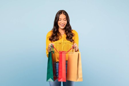 Photo for Delighted young european woman in yellow blouse looking at colorful shopping bags, enjoying retail therapy on light blue background - Royalty Free Image