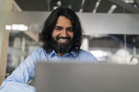 Téléchargez les photos : Portrait rapproché d'un jeune homme barbu indien souriant travaillant sur un ordinateur portable, heureux employé Millénaire de l'Est utilisant l'ordinateur pour le travail ou la communication en ligne, regardant l'écran, Copier l'espace - en image libre de droit