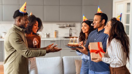 Foto de Grupo alegre de diversos amigos dando pastel de cumpleaños a un chico con sombrero festivo, celebrando bday y haciendo fiesta sorpresa en el interior del apartamento moderno, presentando regalos en la sala de estar. Panorama - Imagen libre de derechos