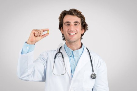 Foto de Alegre doctor masculino sosteniendo la cápsula amarilla, simbolizando consejo médico en suplementos, en bata blanca del laboratorio con el estetoscopio, contra fondo gris - Imagen libre de derechos