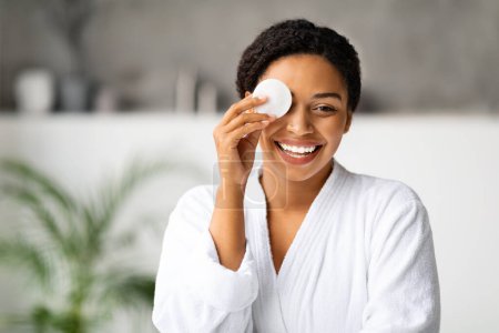Téléchargez les photos : Joyeuse femme noire démaquillant, couvrant l'œil avec du coton et souriant à la caméra, gros plan de femme afro-américaine gaie posant dans la salle de bain tout en faisant la routine de beauté à la maison - en image libre de droit