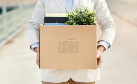 Photo for A person holding a cardboard box filled with office supplies and a green plant, possibly indicating a job change or a new beginning at the workplace, close up, cropped - Royalty Free Image