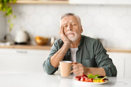 Téléchargez les photos : Homme âgé fatigué reposant la tête sur la main, s'endormant pendant le petit déjeuner, homme âgé dépeignant un moment de fatigue ou de détente, assis à la table de la cuisine dans l'environnement de la maison, espace de copie - en image libre de droit