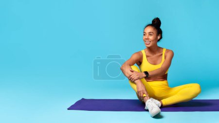 Photo for Young athletic african american woman enjoying sporty healthy lifestyle, sitting on yoga mat, wearing earphones and listening to music with earbuds over blue background, panorama, copy space - Royalty Free Image