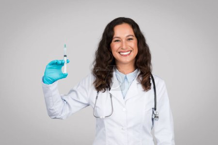 Foto de Doctora confiada con sonrisa alegre sosteniendo la jeringa, preparada para administrar la vacuna, usando guantes médicos y una bata blanca, fondo gris - Imagen libre de derechos