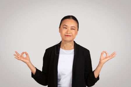 Happy young asian woman in suit with closed eyes meditation, enjoy peace, calm, isolated on gray background, studio. Zen, relax at work, breathing exercises, positive lifestyle, health care