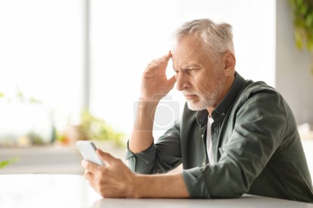 Gestresster älterer Mann mit Kopfschmerzen beim Blick auf das Smartphone zu Hause, besorgter älterer Herr, der beunruhigende Nachrichten liest oder komplexe Apps navigiert, am Tisch in der Küche sitzt, Kopierraum