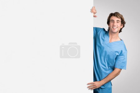 Cheerful male nurse in blue scrubs leaning over blank white board, offering space for medical text or graphics, with friendly smile, banner