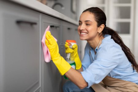 Foto de Mujer contenta con camisa azul y guantes amarillos limpia cuidadosamente los gabinetes de la cocina con tela rosa y spray, sonriendo mientras limpia - Imagen libre de derechos