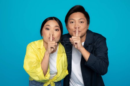 Two playful Asian millennial individuals are gesturing silence with fingers over lips, sharing a secret or quiet moment, against a striking turquoise blue studio backdrop
